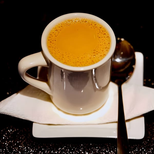 A white mug of frothy, orange-brown Indian chai tea with a spoon on a white saucer.