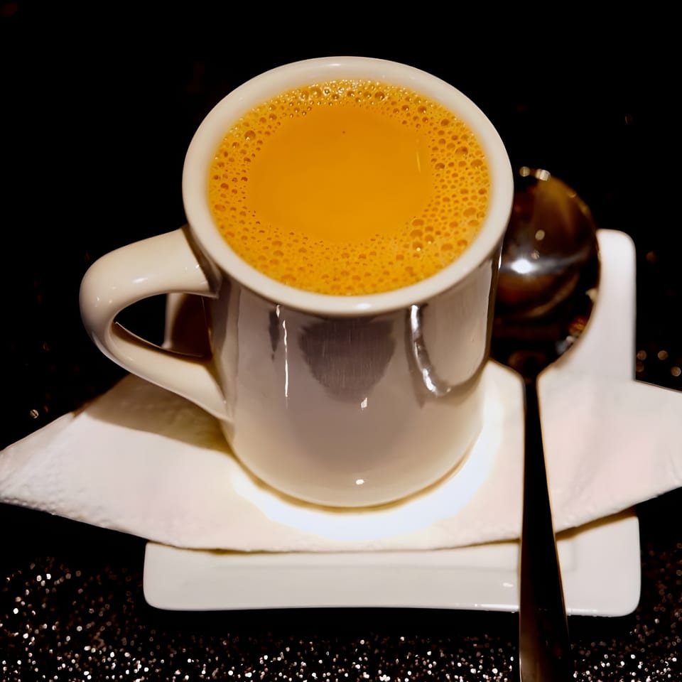 A white mug of frothy, orange-brown Indian chai tea with a spoon on a white saucer.