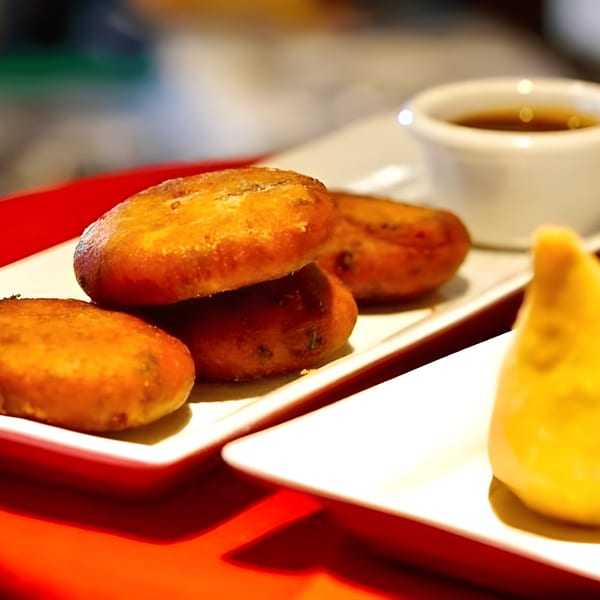 Golden-brown Indian potato patties and a samosa on white plates, with a small cup of sauce.